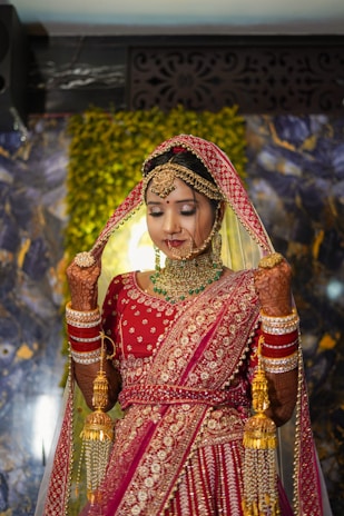 A bride wearing a traditional red and gold embellished lehenga with intricate jewelry, including a heavy necklace and earrings. Her hands are adorned with mehndi and bangles, and she holds a part of her veil with a serene expression. The background features a decorative panel and greenery.