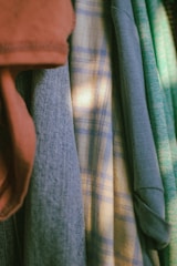 Various articles of clothing are hanging closely together. The garments include a red-brown piece, a blue denim-like fabric, a plaid shirt with light and dark blue checks, and a light green knit sweater. The lighting creates a soft and warm atmosphere.