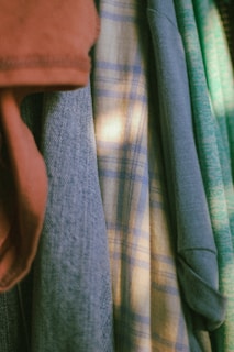 Various articles of clothing are hanging closely together. The garments include a red-brown piece, a blue denim-like fabric, a plaid shirt with light and dark blue checks, and a light green knit sweater. The lighting creates a soft and warm atmosphere.