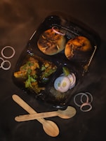 A meal in a black divided container includes two round, grilled pieces of bread garnished with raw onion rings, and a portion of curry with herbs on top. The container is surrounded by additional onion rings and two wooden spoons placed in a crisscross pattern.
