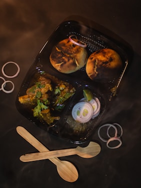 A meal in a black divided container includes two round, grilled pieces of bread garnished with raw onion rings, and a portion of curry with herbs on top. The container is surrounded by additional onion rings and two wooden spoons placed in a crisscross pattern.