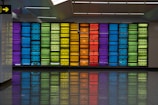 A vibrant wall art display composed of multicolored panels in geometric patterns. The panels are arranged in a grid and feature a variety of colors, including blue, green, yellow, orange, and red. The floor reflects the colors of the panels, adding to the visual impact.
