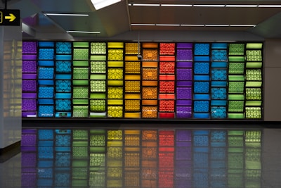A vibrant wall art display composed of multicolored panels in geometric patterns. The panels are arranged in a grid and feature a variety of colors, including blue, green, yellow, orange, and red. The floor reflects the colors of the panels, adding to the visual impact.