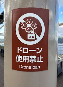 A sign displayed on a pole indicates a ban on drones. The sign features a red circle with a crossed-out drone graphic and text in both Japanese and English stating 'Drone ban'.