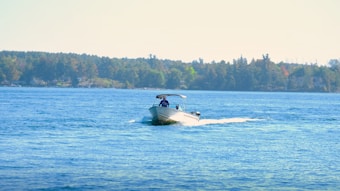 A motorboat glides smoothly across a wide expanse of blue water, creating gentle ripples behind it. A person is seated at the helm, wearing a blue jacket. In the background, a dense line of trees outlines the shoreline under a clear sky.
