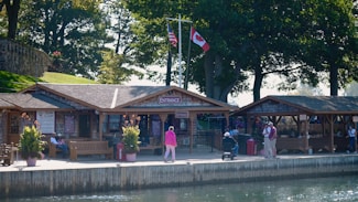 A waterfront scene featuring a wooden boardwalk with rustic buildings labeled as an entrance area. People are sitting and walking, with flags of the United States and Canada displayed. Lush greenery with large trees surrounds the site, creating a peaceful, nature-rich atmosphere.