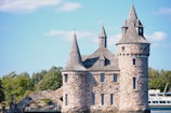 A stone castle-like structure with multiple turrets and conical roofs is set against a backdrop of clear blue skies and surrounded by lush green trees. An arched stone bridge is part of the architecture, and a large boat labeled 'BOAT LINE' is visible on the right side.