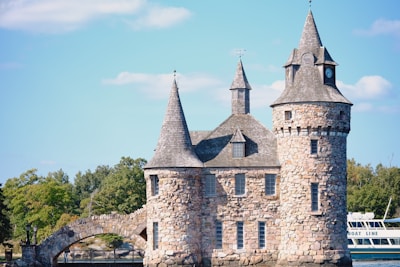 A stone castle-like structure with multiple turrets and conical roofs is set against a backdrop of clear blue skies and surrounded by lush green trees. An arched stone bridge is part of the architecture, and a large boat labeled 'BOAT LINE' is visible on the right side.