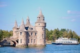 A picturesque stone castle with multiple turrets stands on the edge of a body of water, surrounded by lush greenery. Nearby, a passenger boat labeled 'Rockport Boat Line' traverses the calm waters.