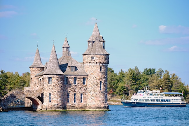 A picturesque stone castle with multiple turrets stands on the edge of a body of water, surrounded by lush greenery. Nearby, a passenger boat labeled 'Rockport Boat Line' traverses the calm waters.