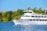 A large white cruise boat named 'Thousand Islander IV' sails on a blue body of water, surrounded by lush green trees. The boat has multiple decks with people enjoying the scenery. The sky is clear and blue, adding to the tranquil setting.