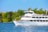 A large white cruise boat named 'Thousand Islander IV' sails on a blue body of water, surrounded by lush green trees. The boat has multiple decks with people enjoying the scenery. The sky is clear and blue, adding to the tranquil setting.