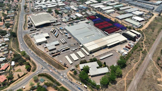 An aerial view of a large industrial area with various warehouses and storage units. There are multiple trucks and shipping containers arranged in sections. Roads and pathways can be seen surrounding the area, and there are trees and patches of greenery scattered around. The surrounding infrastructure and residential buildings edge the industrial zone.
