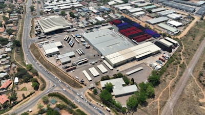 An aerial view of a large industrial area with various warehouses and storage units. There are multiple trucks and shipping containers arranged in sections. Roads and pathways can be seen surrounding the area, and there are trees and patches of greenery scattered around. The surrounding infrastructure and residential buildings edge the industrial zone.