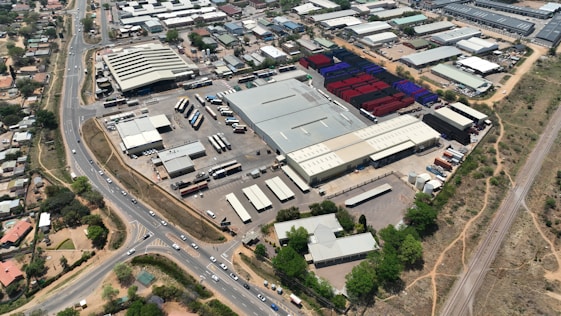 An aerial view of a large industrial area with various warehouses and storage units. There are multiple trucks and shipping containers arranged in sections. Roads and pathways can be seen surrounding the area, and there are trees and patches of greenery scattered around. The surrounding infrastructure and residential buildings edge the industrial zone.