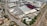An aerial view of a large industrial area with various warehouses and storage units. There are multiple trucks and shipping containers arranged in sections. Roads and pathways can be seen surrounding the area, and there are trees and patches of greenery scattered around. The surrounding infrastructure and residential buildings edge the industrial zone.