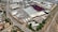 An aerial view of a large industrial area with various warehouses and storage units. There are multiple trucks and shipping containers arranged in sections. Roads and pathways can be seen surrounding the area, and there are trees and patches of greenery scattered around. The surrounding infrastructure and residential buildings edge the industrial zone.