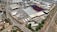 An aerial view of a large industrial area with various warehouses and storage units. There are multiple trucks and shipping containers arranged in sections. Roads and pathways can be seen surrounding the area, and there are trees and patches of greenery scattered around. The surrounding infrastructure and residential buildings edge the industrial zone.