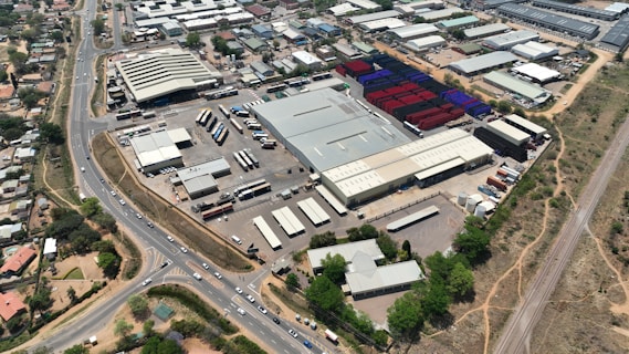 An aerial view of a large industrial area with various warehouses and storage units. There are multiple trucks and shipping containers arranged in sections. Roads and pathways can be seen surrounding the area, and there are trees and patches of greenery scattered around. The surrounding infrastructure and residential buildings edge the industrial zone.