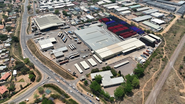 An aerial view of a large industrial area with various warehouses and storage units. There are multiple trucks and shipping containers arranged in sections. Roads and pathways can be seen surrounding the area, and there are trees and patches of greenery scattered around. The surrounding infrastructure and residential buildings edge the industrial zone.