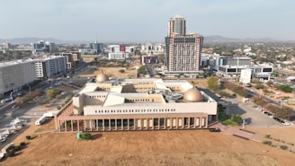 An aerial view of an urban environment with a focus on a modern, low-rise building with domed roofs. Surrounding this structure are taller, more contemporary high-rise buildings, and a network of roads with vehicles. The landscape beyond the city includes distant hills under a clear sky.