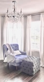 A plush armchair with a soft blue upholstery sits beside a large, tufted ottoman in a room with dark wooden floors. White drapes frame the windows behind, casting a soft light into the space. A crystal chandelier hangs from the ceiling, adding an elegant touch to the cozy setting.