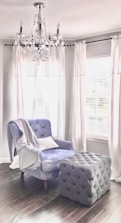 A plush armchair with a soft blue upholstery sits beside a large, tufted ottoman in a room with dark wooden floors. White drapes frame the windows behind, casting a soft light into the space. A crystal chandelier hangs from the ceiling, adding an elegant touch to the cozy setting.