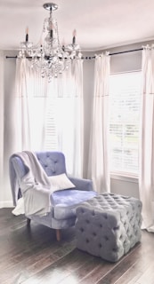 A plush armchair with a soft blue upholstery sits beside a large, tufted ottoman in a room with dark wooden floors. White drapes frame the windows behind, casting a soft light into the space. A crystal chandelier hangs from the ceiling, adding an elegant touch to the cozy setting.