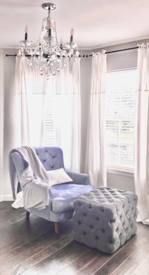 A plush armchair with a soft blue upholstery sits beside a large, tufted ottoman in a room with dark wooden floors. White drapes frame the windows behind, casting a soft light into the space. A crystal chandelier hangs from the ceiling, adding an elegant touch to the cozy setting.