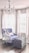 A plush armchair with a soft blue upholstery sits beside a large, tufted ottoman in a room with dark wooden floors. White drapes frame the windows behind, casting a soft light into the space. A crystal chandelier hangs from the ceiling, adding an elegant touch to the cozy setting.