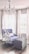 A plush armchair with a soft blue upholstery sits beside a large, tufted ottoman in a room with dark wooden floors. White drapes frame the windows behind, casting a soft light into the space. A crystal chandelier hangs from the ceiling, adding an elegant touch to the cozy setting.