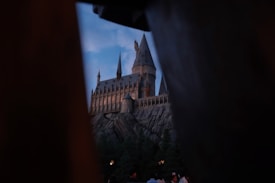 A majestic castle with tall towers and spires is situated on a rocky cliff, surrounded by lush greenery. The sky is dimly lit, suggesting dusk or dawn, and the castle is illuminated from within, creating a mysterious and enchanting atmosphere.