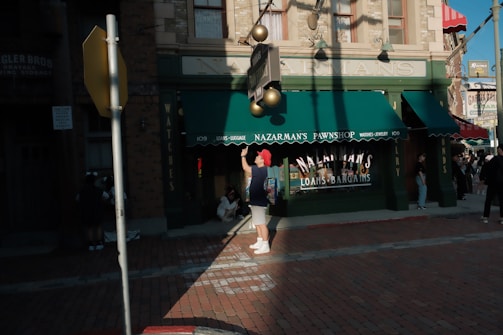 A street scene in front of a building labeled Nazarman's Pawnshop. The shop has green awnings and signage advertising loans and bargains. Several people are present, including someone with a red hat adjusting a light fixture. Shadows are cast across the brick sidewalk in the late afternoon light.