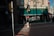 A street scene in front of a building labeled Nazarman's Pawnshop. The shop has green awnings and signage advertising loans and bargains. Several people are present, including someone with a red hat adjusting a light fixture. Shadows are cast across the brick sidewalk in the late afternoon light.