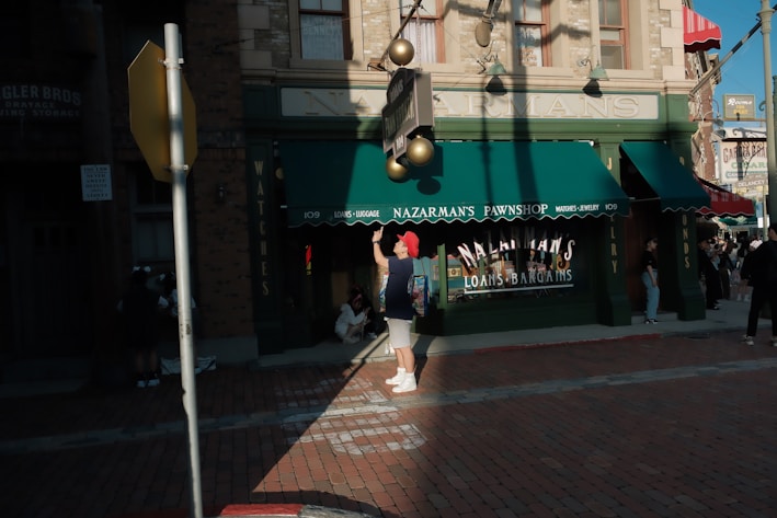A street scene in front of a building labeled Nazarman's Pawnshop. The shop has green awnings and signage advertising loans and bargains. Several people are present, including someone with a red hat adjusting a light fixture. Shadows are cast across the brick sidewalk in the late afternoon light.