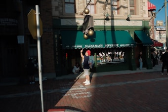 A street scene in front of a building labeled Nazarman's Pawnshop. The shop has green awnings and signage advertising loans and bargains. Several people are present, including someone with a red hat adjusting a light fixture. Shadows are cast across the brick sidewalk in the late afternoon light.