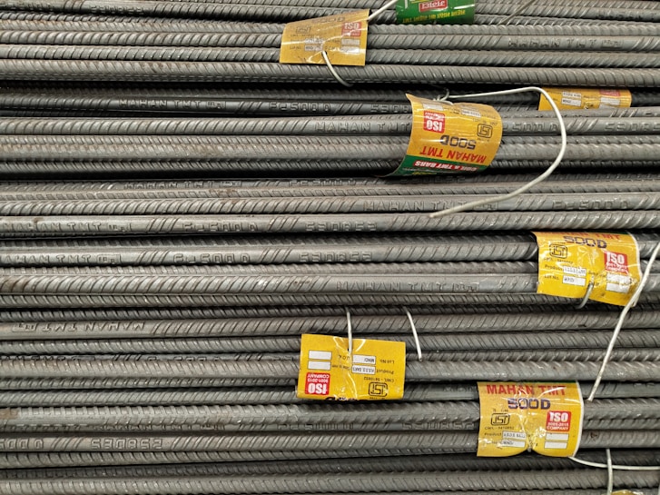 A stack of steel rebars neatly arranged with yellow labels attached to several bars. The labels contain various specifications and certifications. The rebars have a textured surface, indicating they are designed for reinforced concrete use.