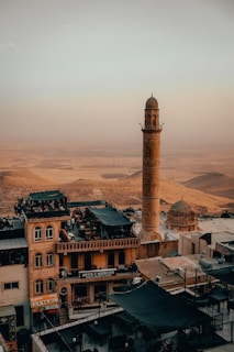 A tall minaret and dome of a mosque stand prominently in a scenic hillside town. Several buildings in traditional architectural style are visible, with some rooftops having seating areas. The landscape stretches into an arid expanse in the background, evoking a sense of cultural and geographical connection.