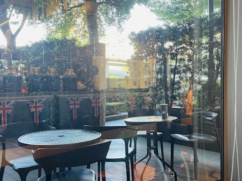 An outdoor seating area with round tables and black chairs, surrounded by greenery. Union Jack flags are displayed on the wall behind, and there are some decorative jars or pots on the ledge. Sunlight filters through the trees, creating a calm and inviting atmosphere.