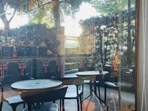 An outdoor seating area with round tables and black chairs, surrounded by greenery. Union Jack flags are displayed on the wall behind, and there are some decorative jars or pots on the ledge. Sunlight filters through the trees, creating a calm and inviting atmosphere.