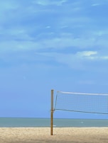 A sandy beach with a volleyball net in the foreground, under a clear blue sky. The ocean is visible in the background, creating a serene and open atmosphere.