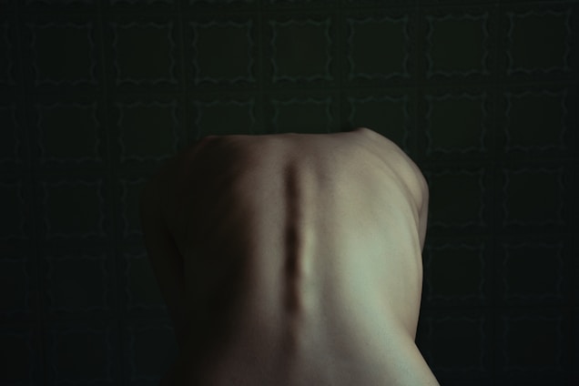 A dimly lit image focusing on the upper back of a human figure in a seated position, against a dark green patterned background. The lighting accentuates the spine and shoulder blades.