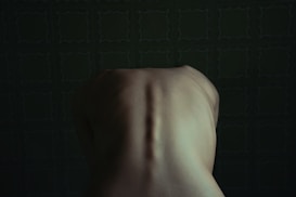 A dimly lit image focusing on the upper back of a human figure in a seated position, against a dark green patterned background. The lighting accentuates the spine and shoulder blades.