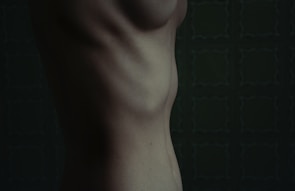 A close-up of a human torso viewed from the side, showcasing the contours of the ribcage and abdomen against a dark, patterned background.