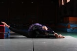 A gymnast performs a stretching exercise on a mat in a dimly lit gymnasium. The athlete is wearing a purple gymnastics leotard with sparkly details, and is in a deep split position with their torso arched backwards. Soft mats are visible in the background.