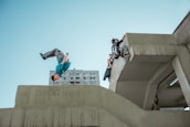 A person is performing a backflip off a concrete structure while another person climbs over a ledge. In the background, there is a high-rise building. The sky is clear, and the scene captures a dynamic moment of urban parkour.