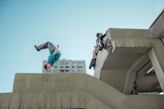 A person is performing a backflip off a concrete structure while another person climbs over a ledge. In the background, there is a high-rise building. The sky is clear, and the scene captures a dynamic moment of urban parkour.