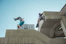 A person is performing a backflip off a concrete structure while another person climbs over a ledge. In the background, there is a high-rise building. The sky is clear, and the scene captures a dynamic moment of urban parkour.