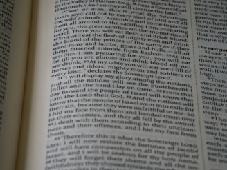 The image displays a close-up of a book page with printed text. The text is formatted in columns with chapter and verse numbers, suggesting it is from a religious or historical document. The pages show signs of use, with slight shadows and lighting focusing on certain lines.