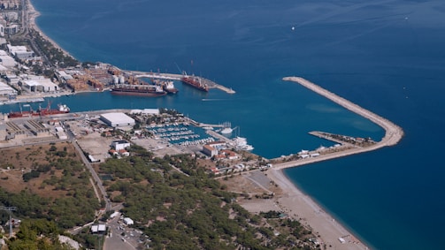 Aerial view of a coastal port with several ships docked. The area features a marina with numerous smaller boats, surrounded by industrial facilities. Further inland, green vegetation and urban buildings are visible. The coastline curves gently, bordered by clear blue waters.